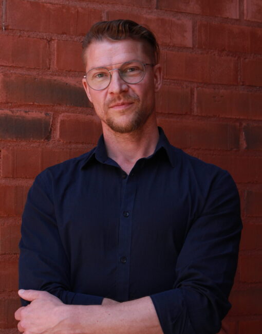 Muotokuva miehestä, jolla on pyöreät metallikehyksiset silmälasit, tummansininen kauluspaita ja lyhyt parta, seisomassa punatiiliseinää vasten. Valo on pehmeää ja tasaista.